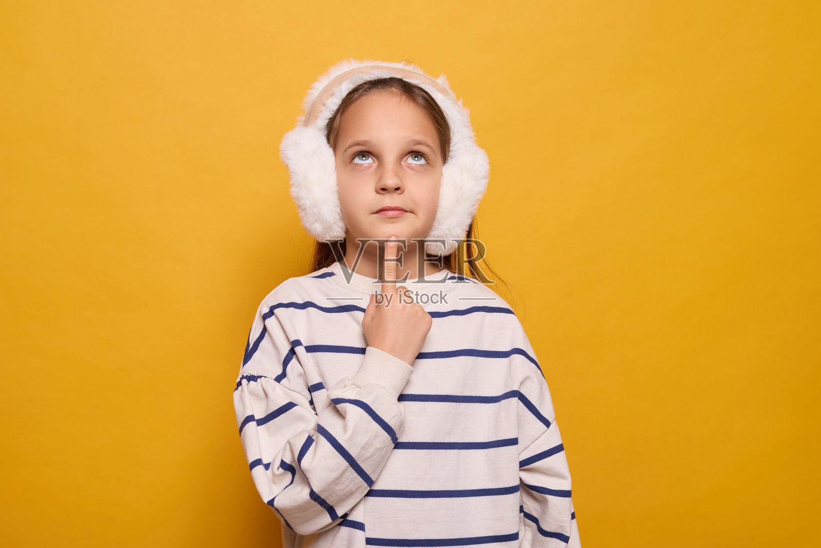 一个女孩戴着毛茸茸的耳罩，穿着条纹上衣，若有所思地抚摸着下巴。她的表情暗示着她在思考一个想法或在鲜艳的黄色背景下做白日梦。照片摄影图片