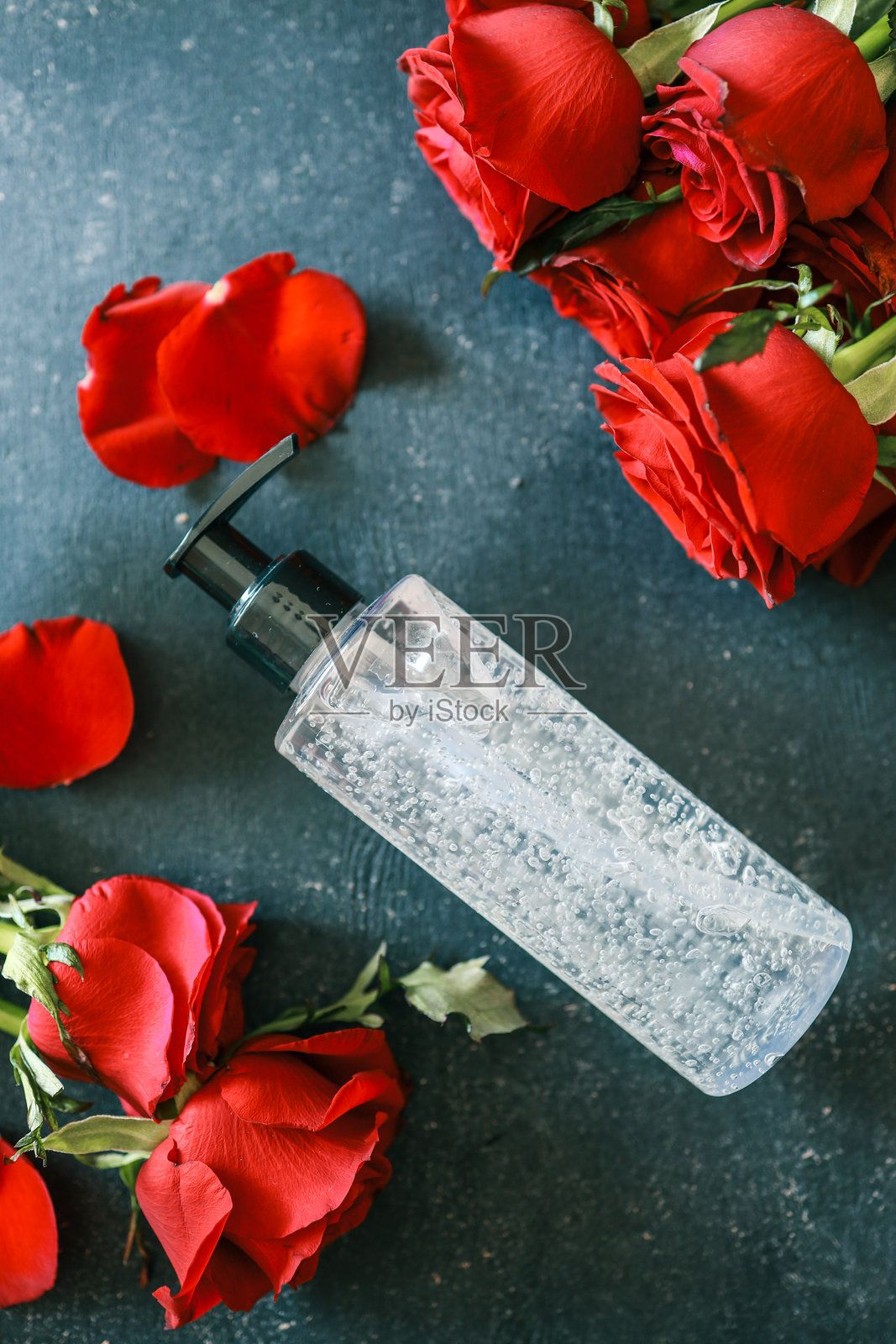 透明泵瓶装有清澈的凝胶，置于鲜艳的红玫瑰和花瓣之间，背景为深色纹理表面，突显现代和清新的美感。照片摄影图片