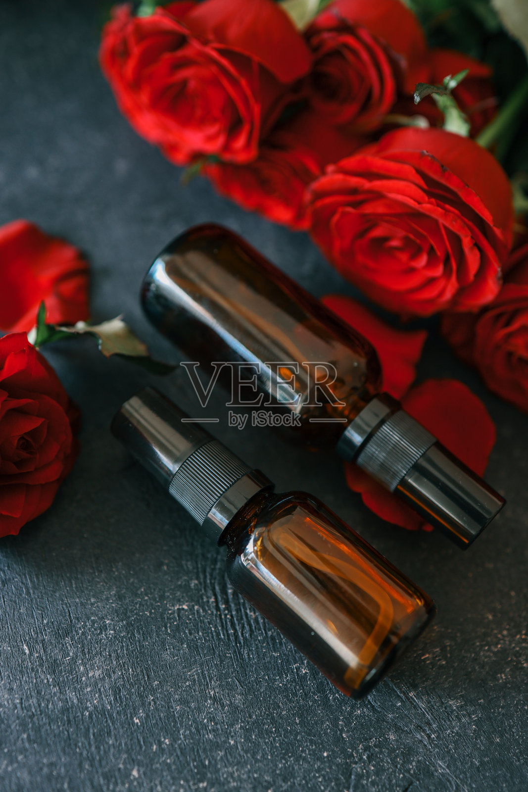 琥珀色喷雾瓶配有黑色喷嘴，放置在深色纹理表面上，周围环绕着鲜艳的红玫瑰。粗犷的木质背景增添了温暖自然的氛围。照片摄影图片