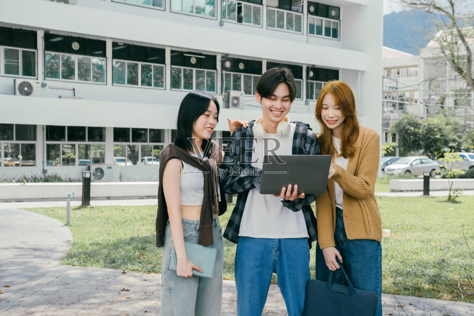 三名快乐的年轻大学生在大学建筑外一起使用笔记本电脑，享受着宜人的天气和合作学习的乐趣。照片摄影图片