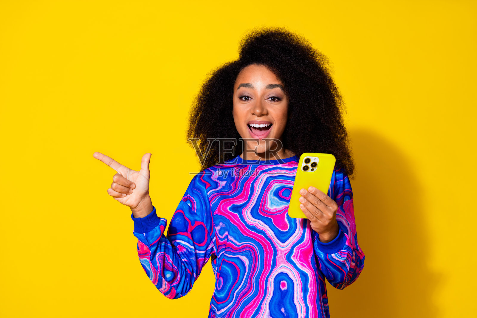 可爱的年轻女士兴奋地指着空白处，手持设备，穿着时尚的彩色蓝色服装，背景为黄色。照片摄影图片