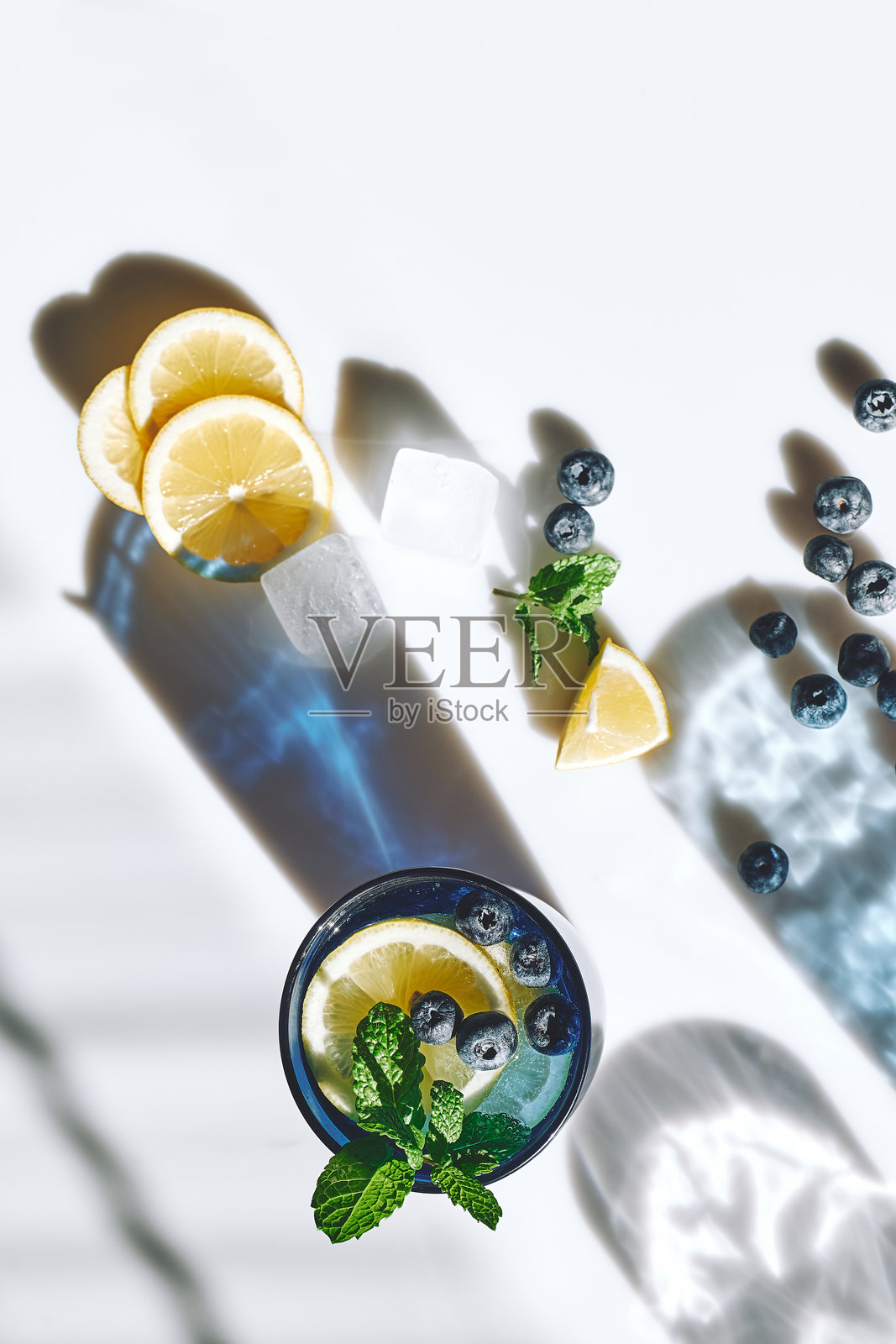 夏季清爽的蓝莓薄荷冰柠檬饮料，配有冰块和柠檬片，摆放在白色表面上，投下阴影。新鲜健康的冷柠檬饮品。柠檬浸泡水。照片摄影图片
