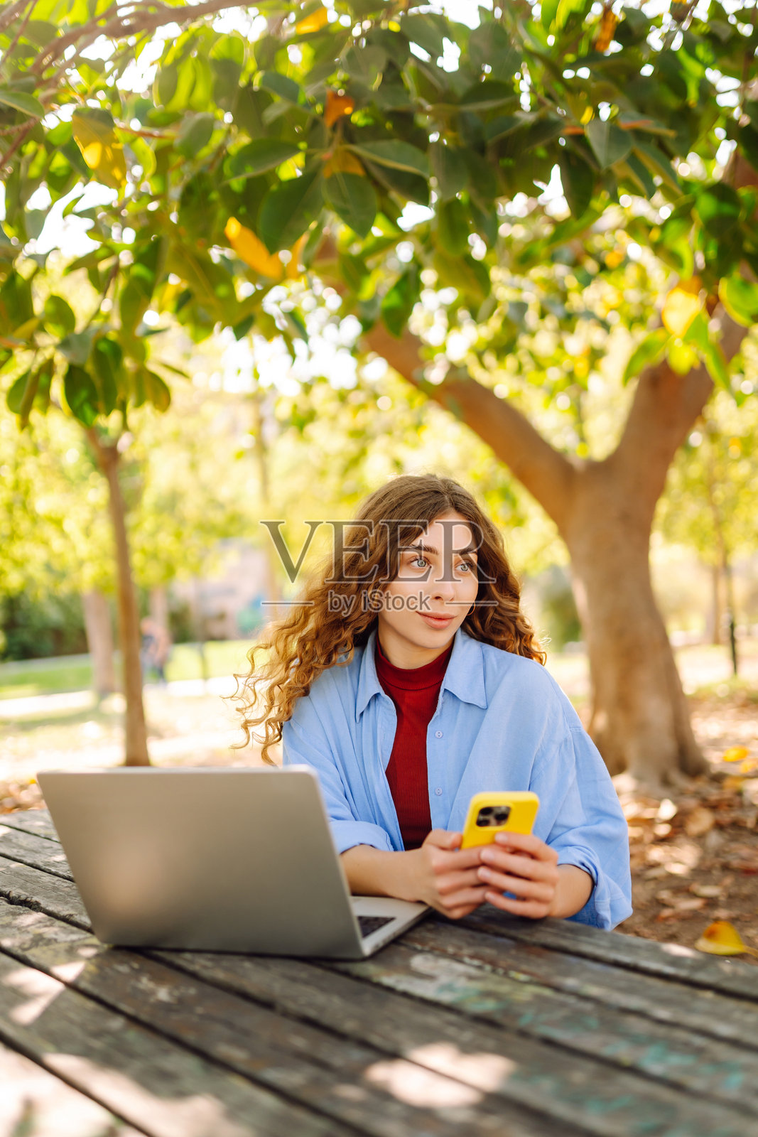 在湖边的户外咖啡馆里，一位快乐的女性坐在桌旁，手里拿着笔记本电脑和手机。照片摄影图片
