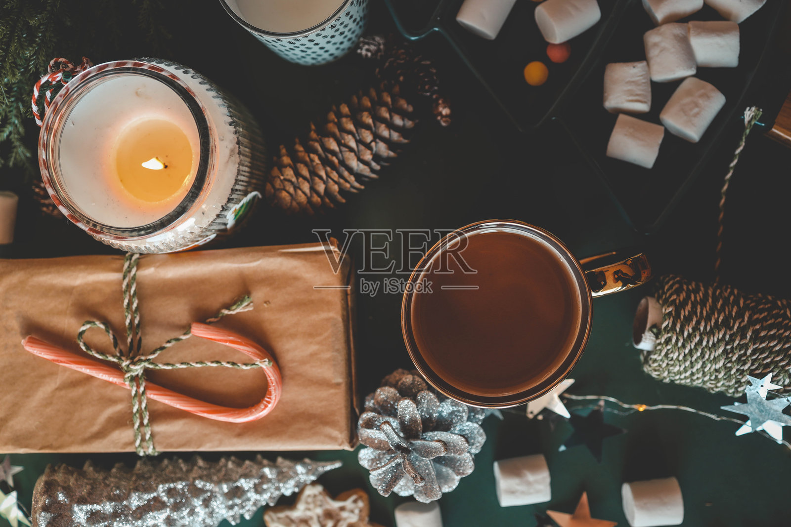 从上方俯视一个节日餐桌，上面有一杯茶、闪烁的蜡烛、松果、棉花糖和装饰元素，背景为绿色，营造出温馨的圣诞氛围。照片摄影图片