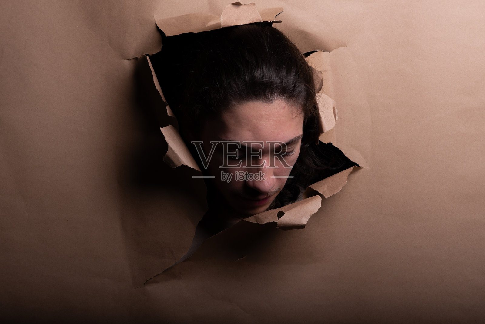 男人的脸和头从棕色纸张的一个洞里探出来，双手推开纸张。照片摄影图片