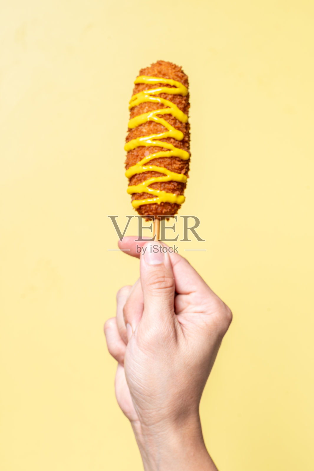 一只手握着淋有芥末酱的酥脆韩国玉米热狗，背景为纯黄色。照片摄影图片