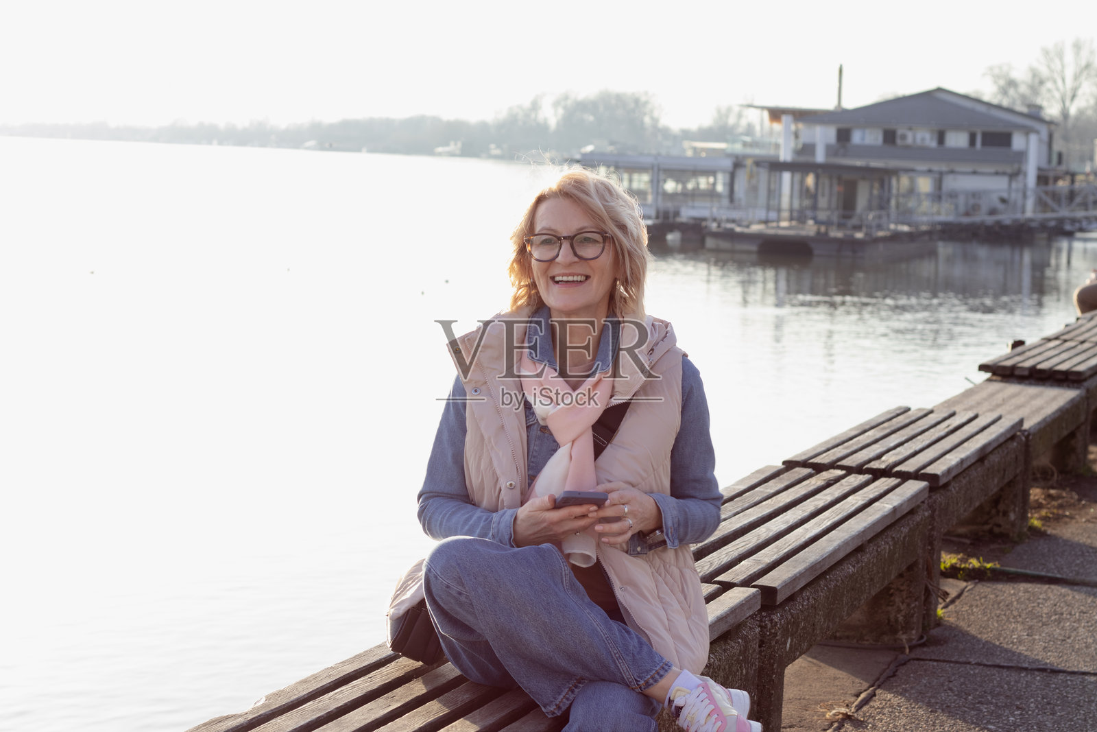 成熟的金发女性戴着眼镜，坐在河边的长椅上，正在用手机发信息。照片摄影图片