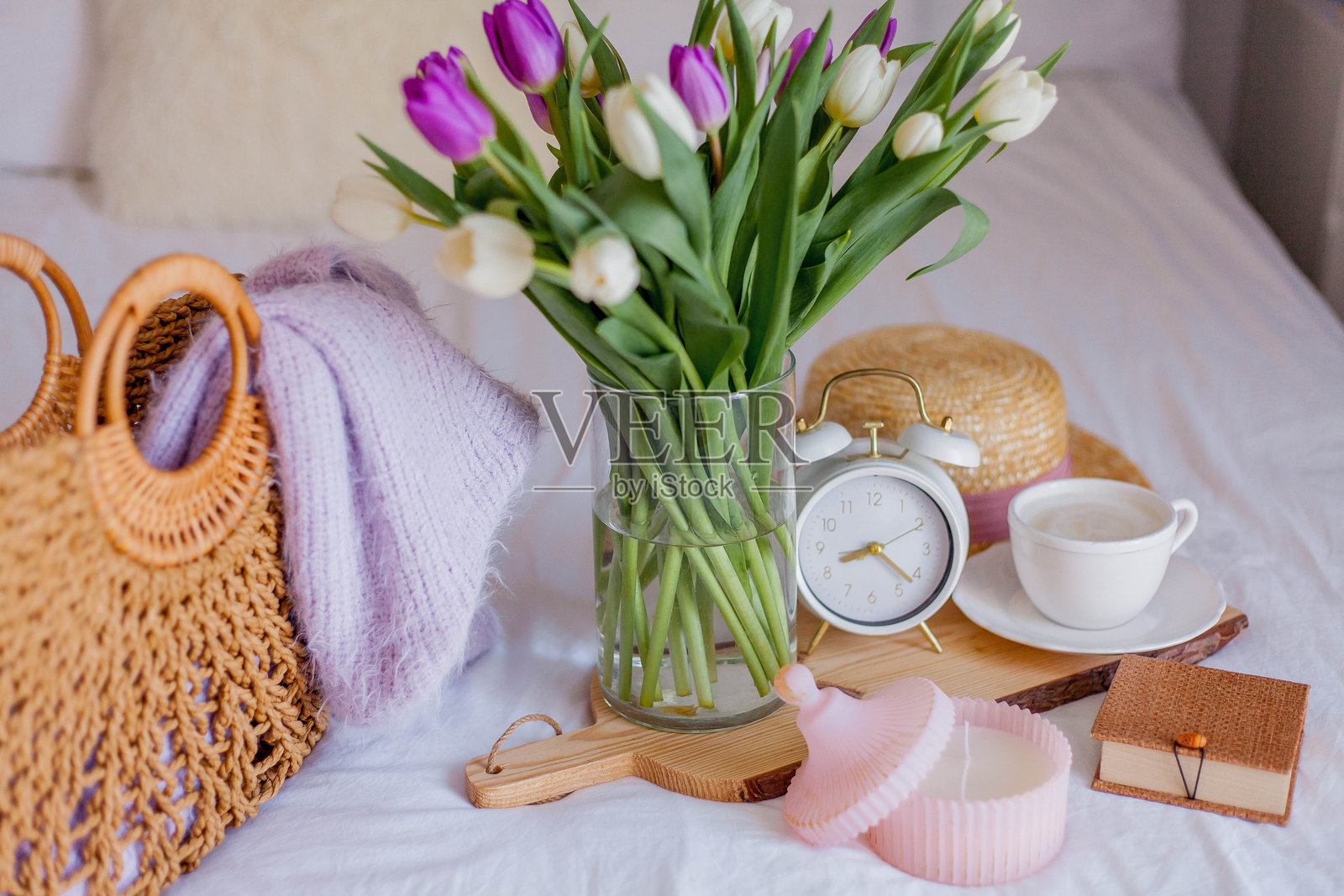 一束郁金香放在玻璃花瓶中，旁边有一个白色的闹钟、一支蜡烛、一张便条和一个卡布奇诺杯，摆放在床上。这是女性的物品。床上的早餐。照片摄影图片
