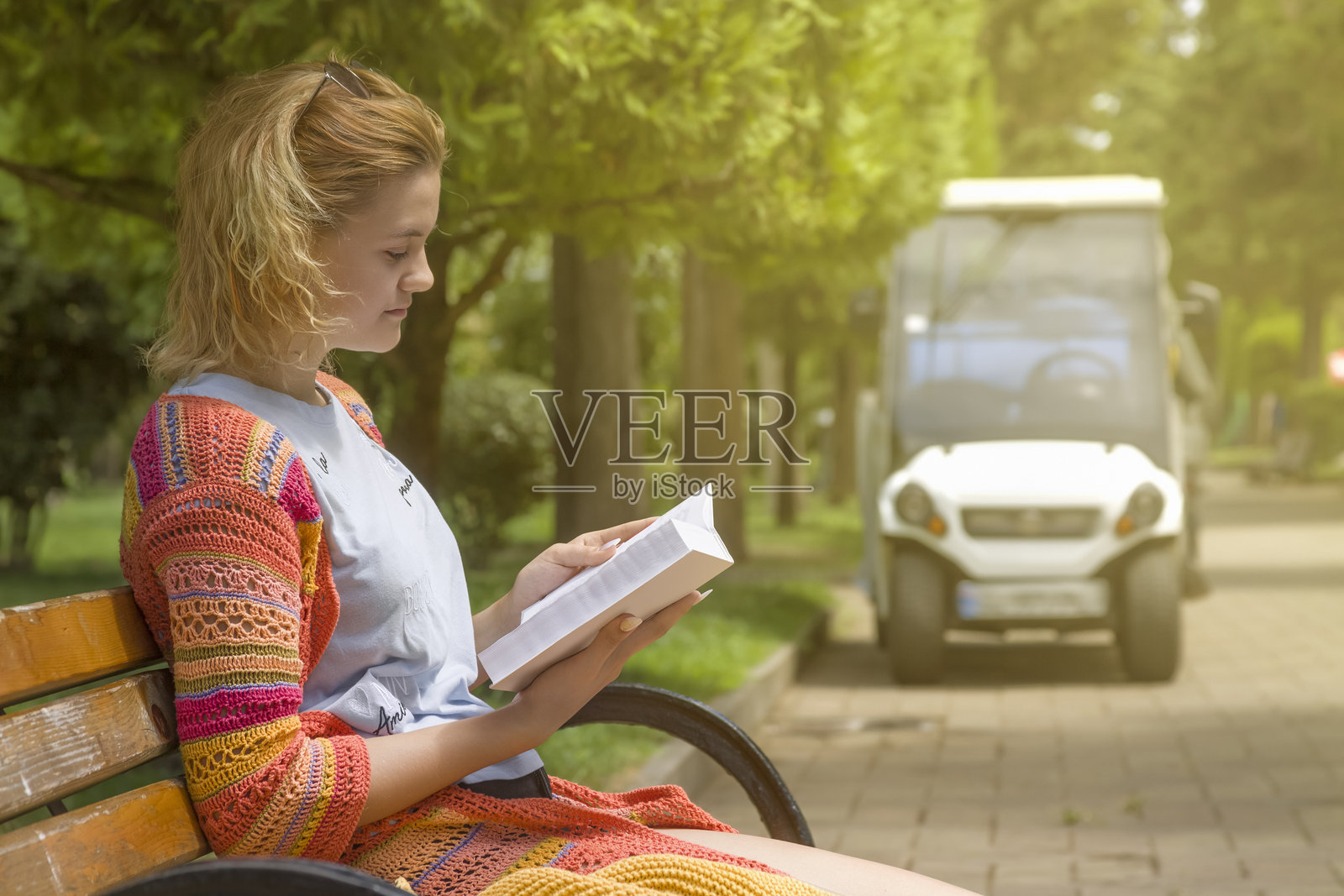 一个穿着棉质针织衣服的小女孩坐在城市公园的木长椅上，享受着温暖的夏日，专心阅读一本书。特写镜头。照片摄影图片