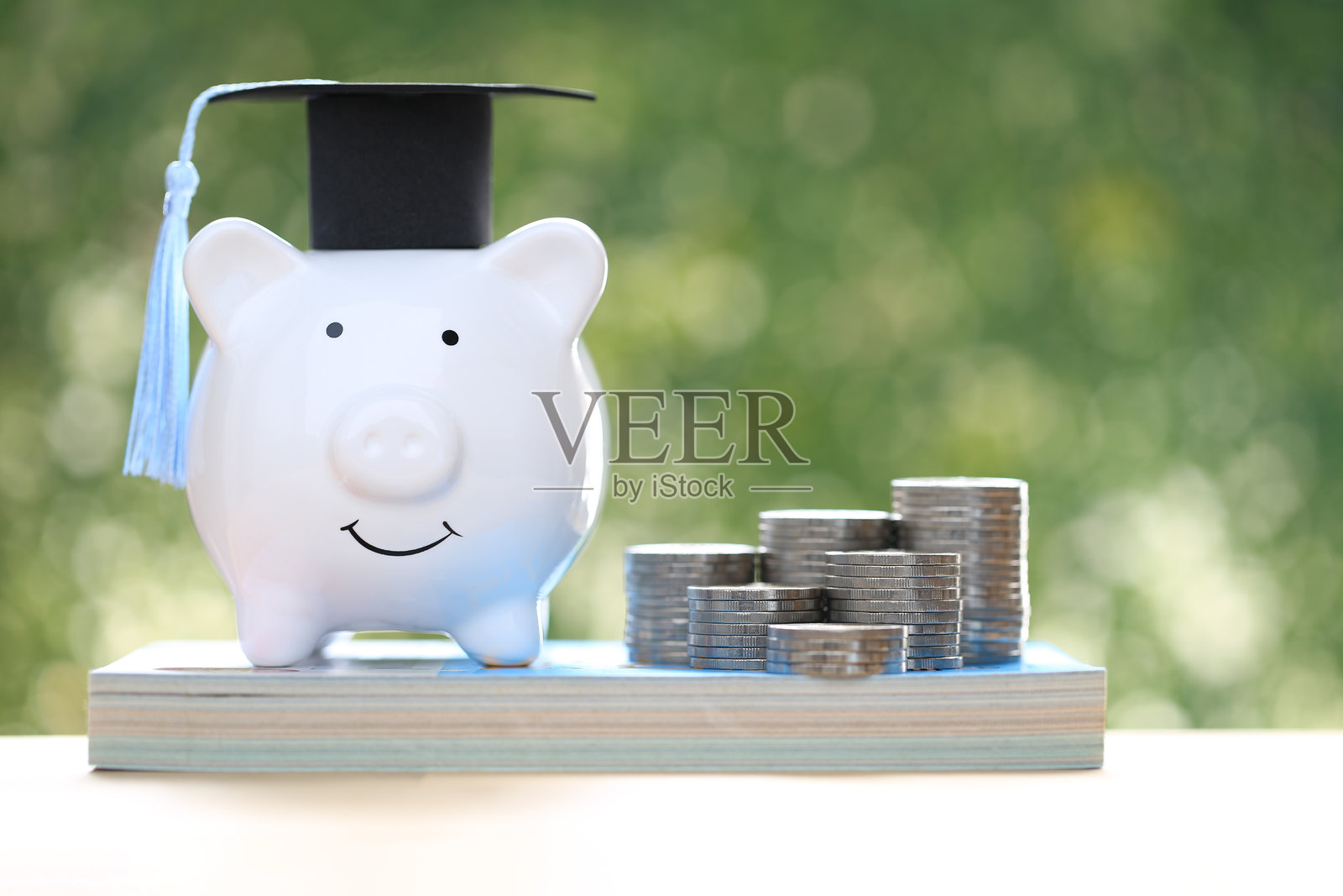 毕业帽放在储蓄罐上，旁边有一堆硬币和钞票，背景为自然绿色，寓意为教育储蓄。照片摄影图片