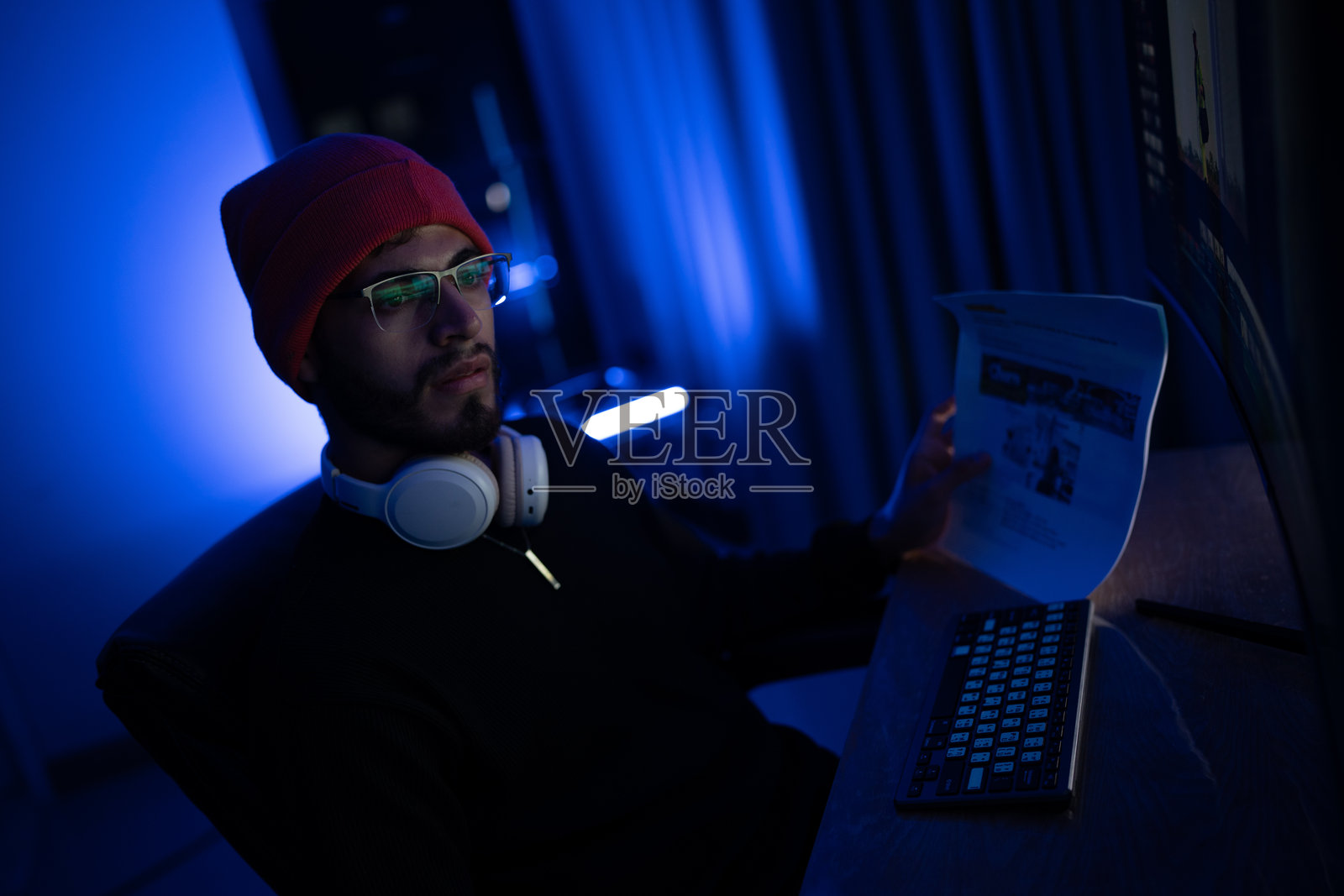 年轻男子戴着耳机在昏暗的房间里使用电脑，夜晚的蓝色灯光映照着他。照片摄影图片