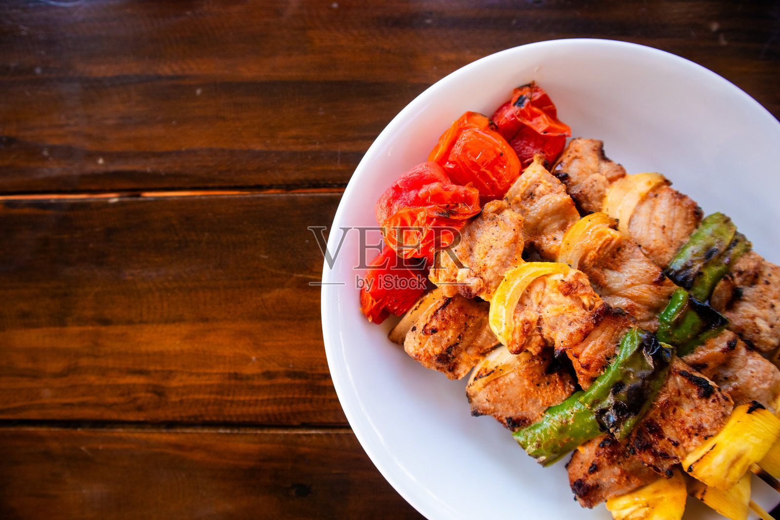 白色盘子里的烧烤串放在木桌上。烧烤猪肉是一种烹饪方法，肉类、蔬菜、海鲜等被烤制或熏制。照片摄影图片