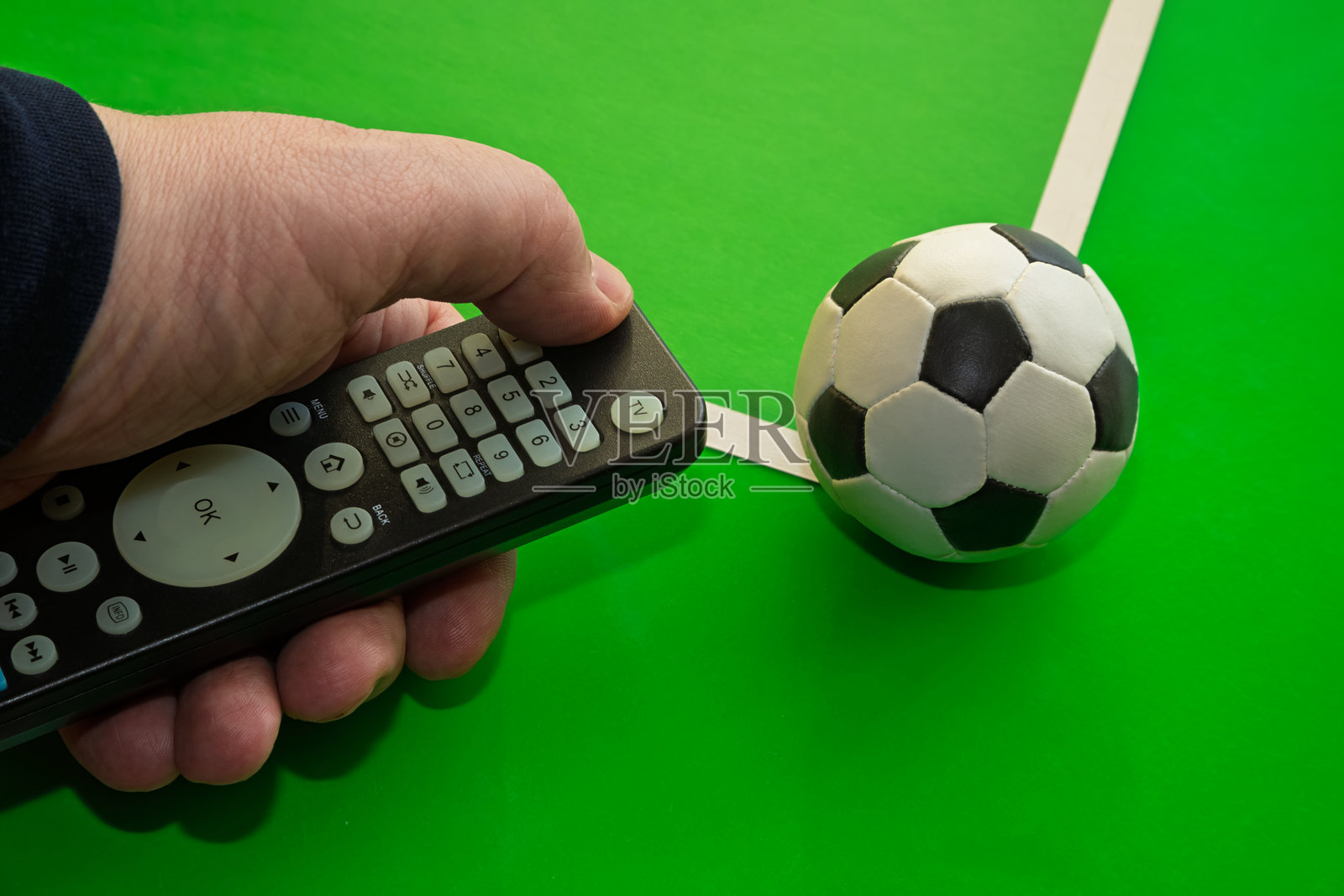 绿色背景的足球场，近距离拍摄的足球和手按下电视遥控器的按钮，非常适合足球周末、现场体育直播、电视上的足球和体育博彩。照片摄影图片