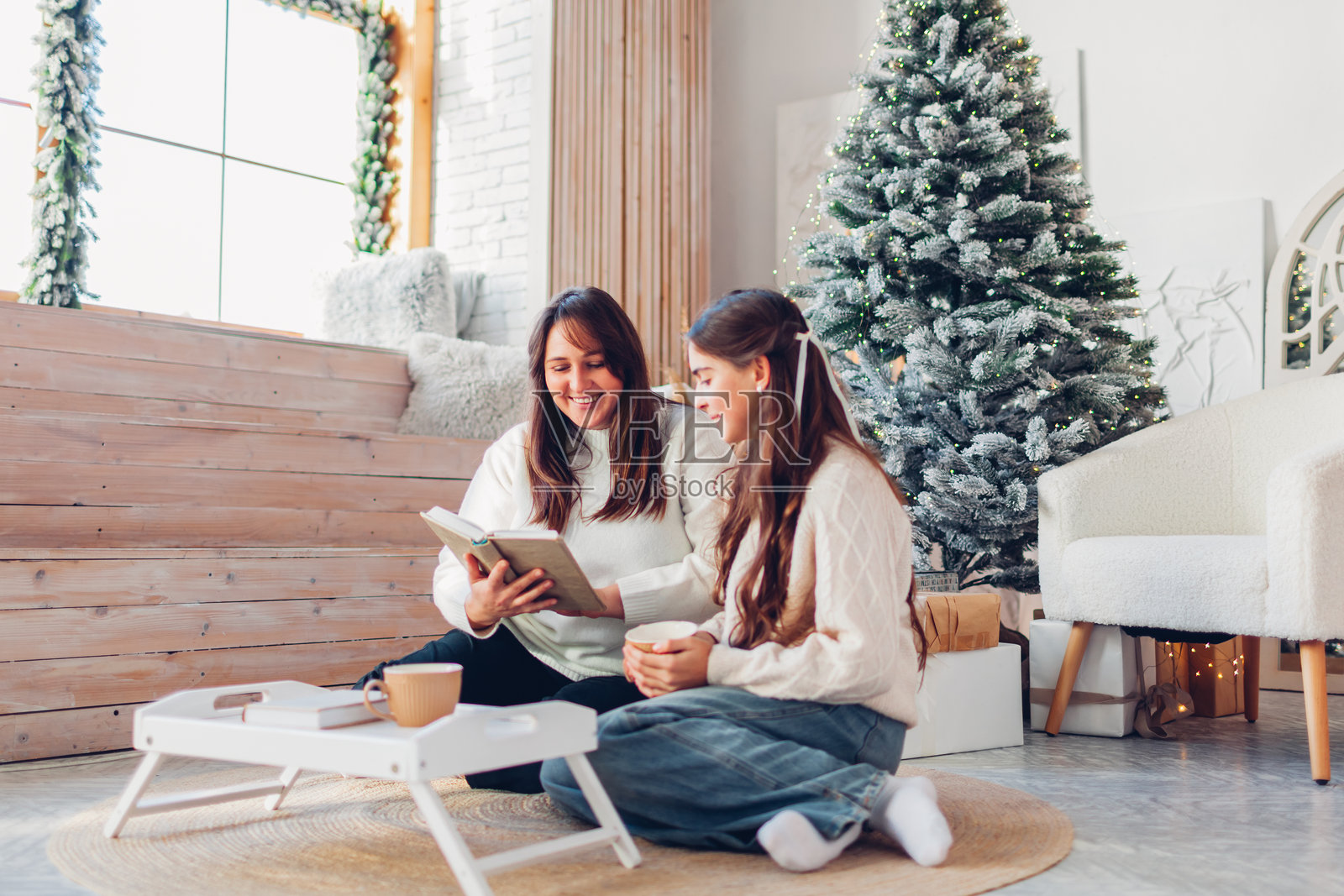 母亲和她的青少年女儿在家里的圣诞树旁放松地阅读书籍。全家人坐在地板上喝茶。照片摄影图片
