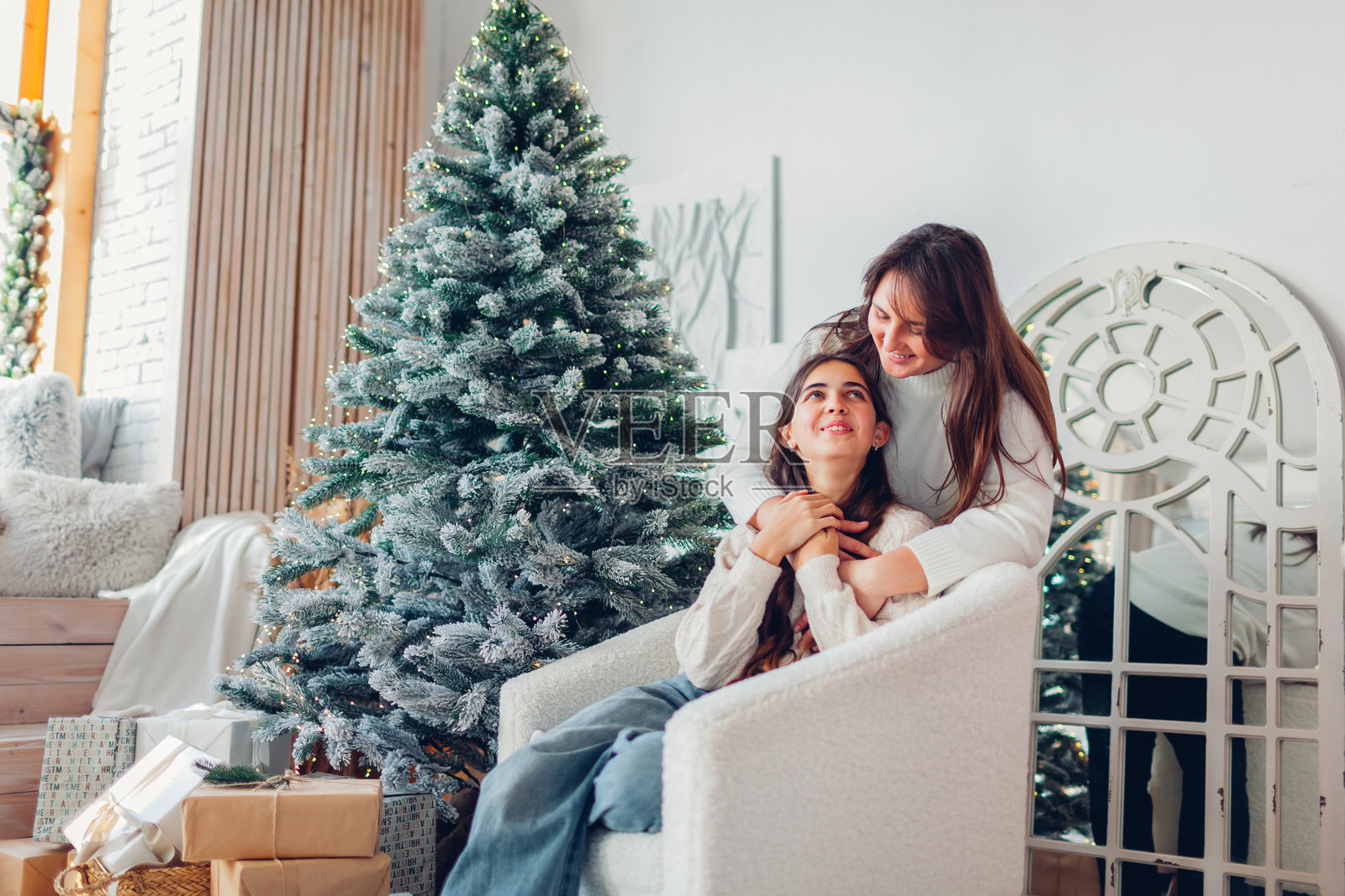 快乐的母亲在装饰好的圣诞树旁拥抱她的青少年女儿。家庭庆祝节日。照片摄影图片