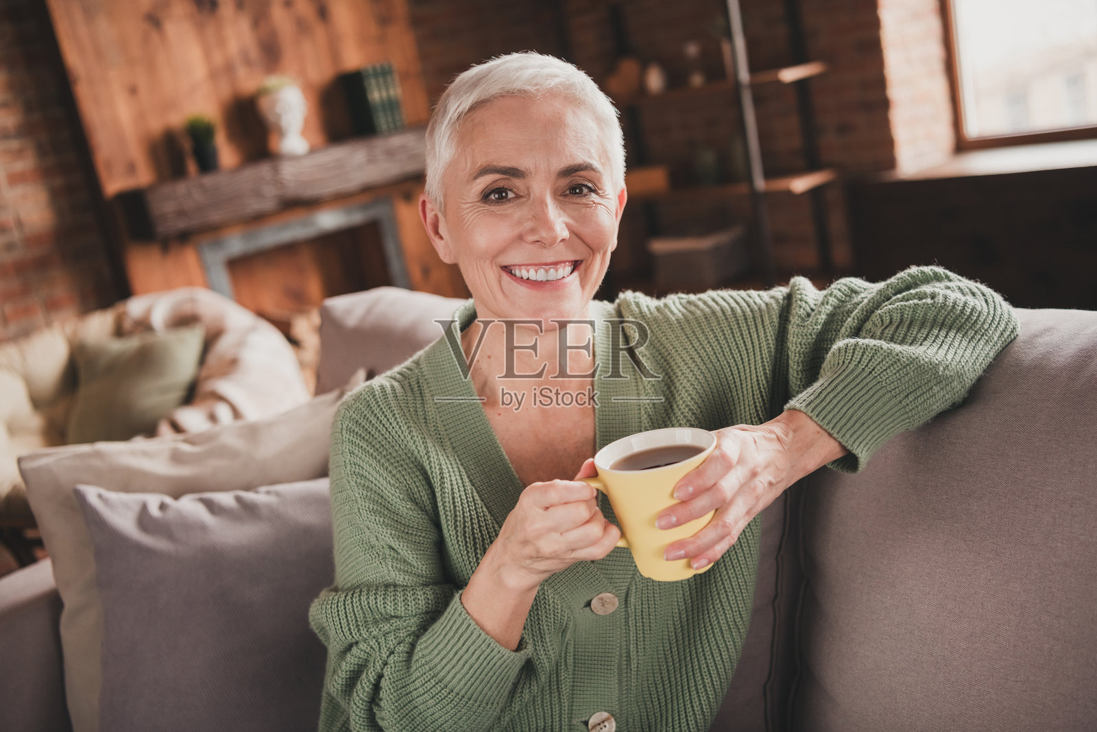 迷人的老年女性在家中悠闲地享受周末，穿着时尚的绿色毛衣，手中捧着温暖的饮品。照片摄影图片