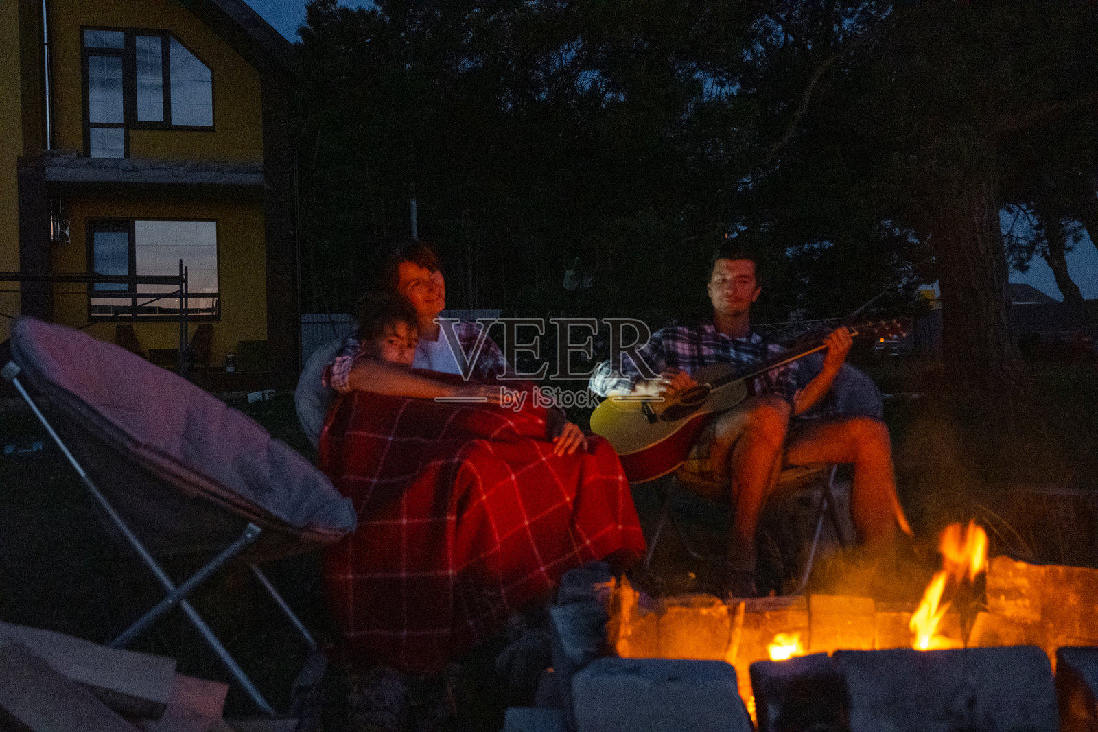 一家人围坐在篝火旁，一名男子弹着吉他，一名女子在旁倾听并跟着唱，我的女儿裹着毯子。家庭夏季的傍晚，在院子里。照片摄影图片