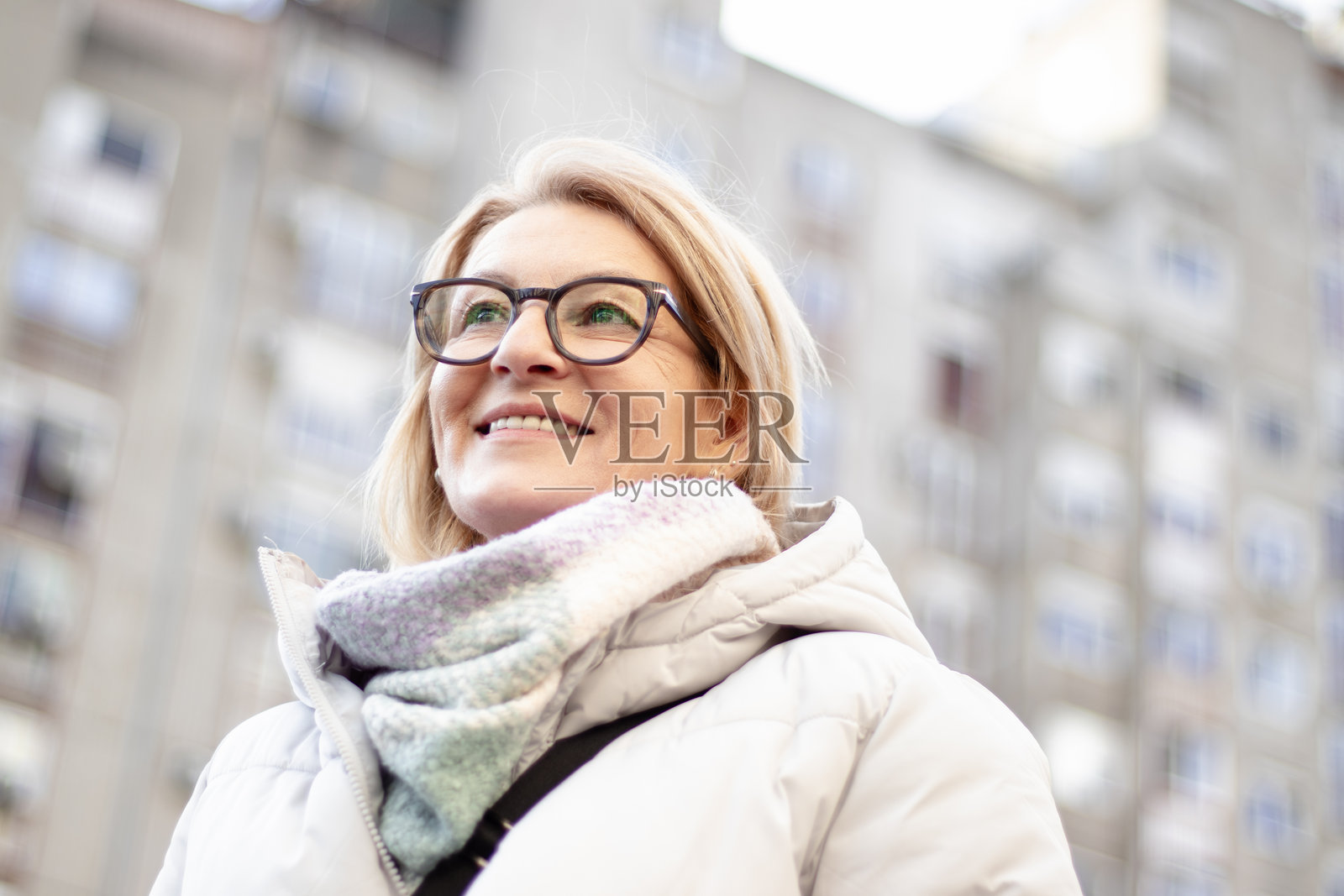 一位成熟的金发女性戴着眼镜，站在冬日的建筑前。照片摄影图片