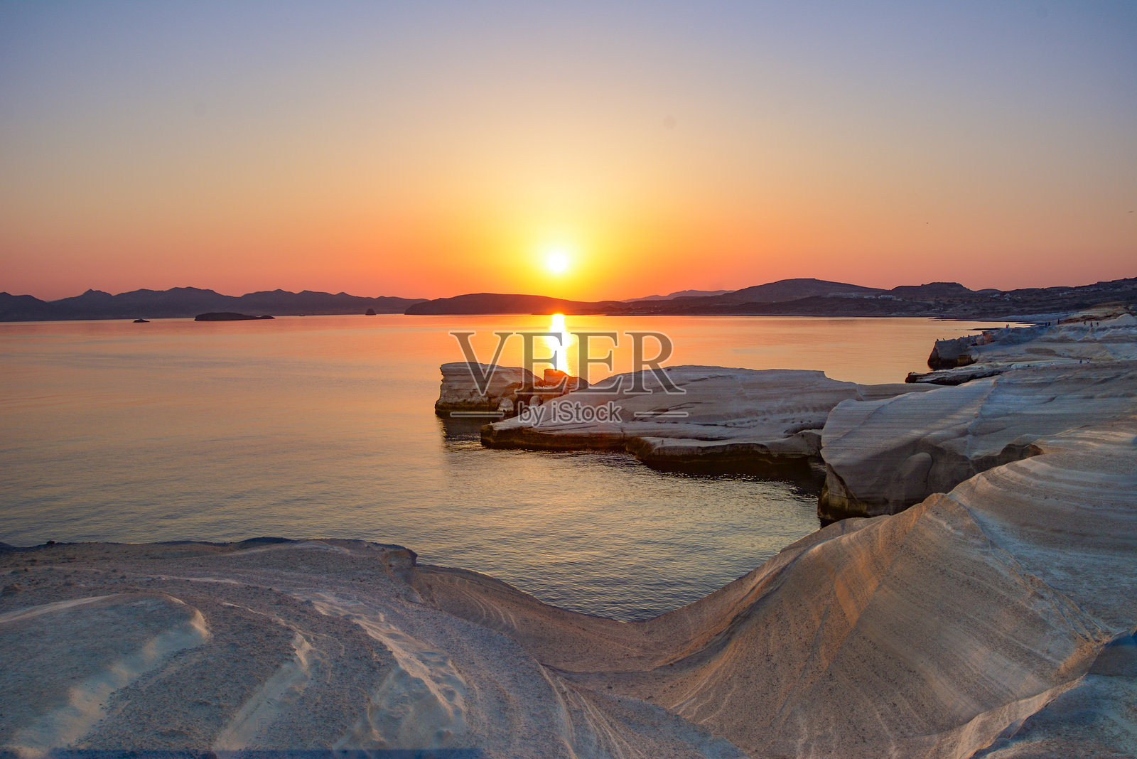 米洛斯岛萨拉基尼科海滩的日出，橙色的天空映衬着白色悬崖、岩石和蔚蓝的爱琴海海岸线。照片摄影图片