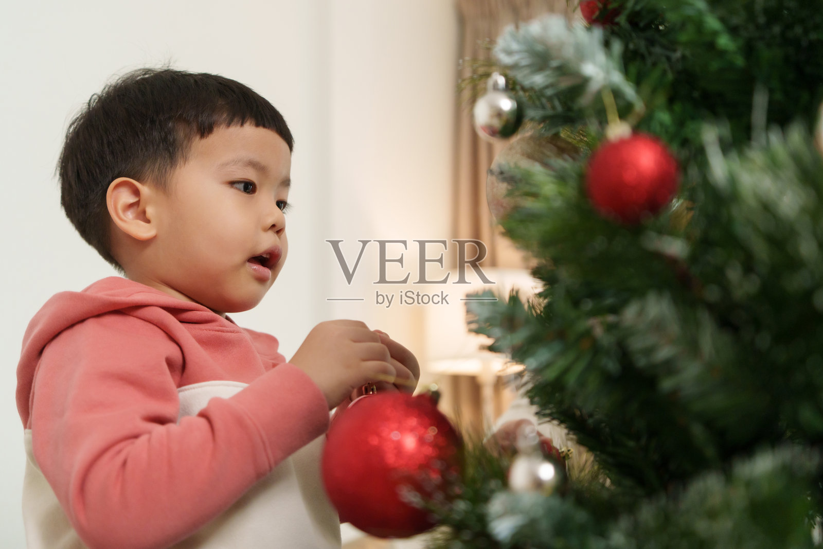 亚洲小孩小心翼翼地将红色装饰挂在圣诞树上，周围是一个温馨宜人的客厅，洋溢着节日的欢乐气氛。柔和的灯光和装饰品增添了节日的氛围。照片摄影图片