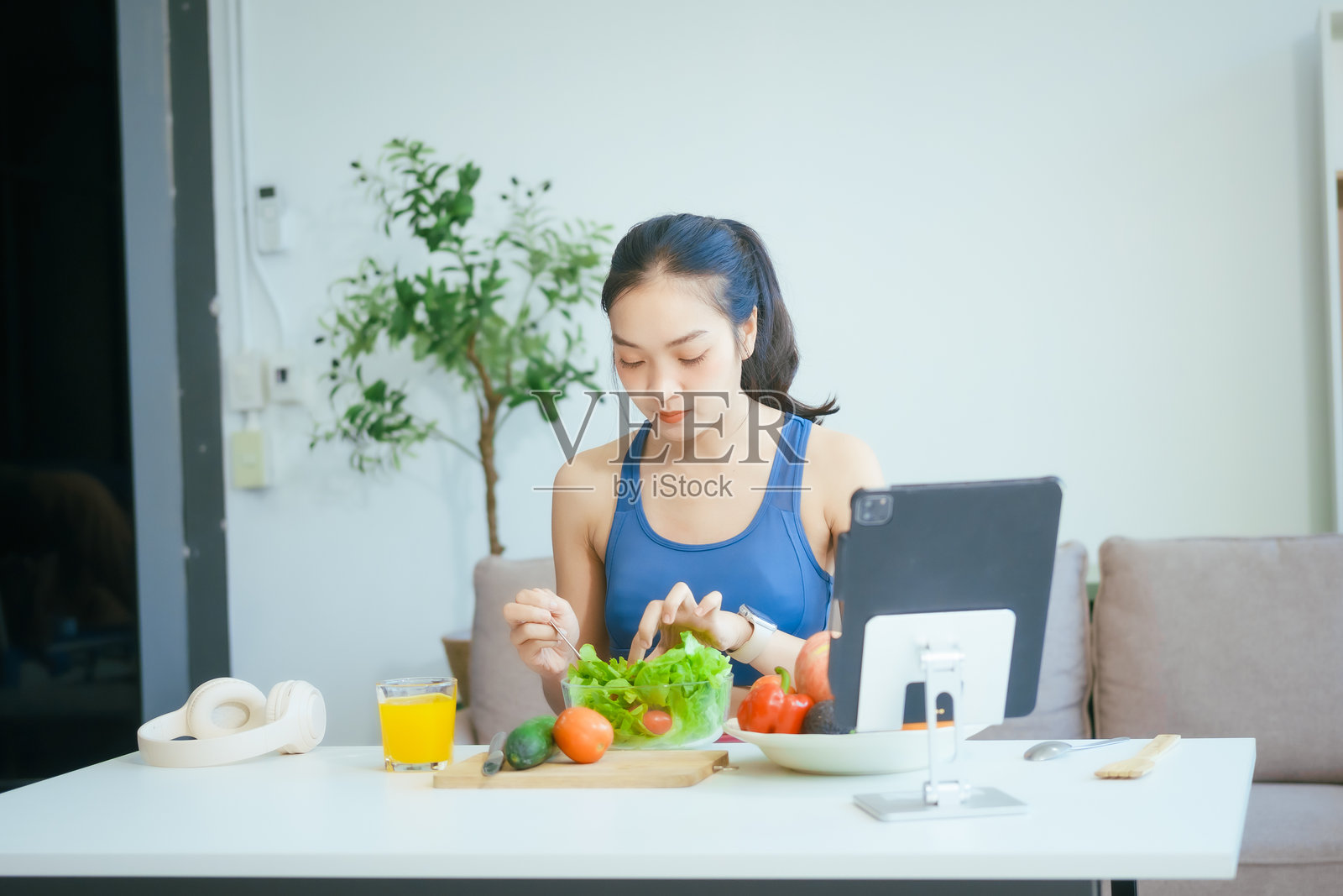 一位快乐的亚洲女性穿着运动服，坐在沙发上，享受着一碗沙拉、水果、新鲜蔬菜、橙汁和健身补剂，刚刚完成了一次家庭健身锻炼。照片摄影图片