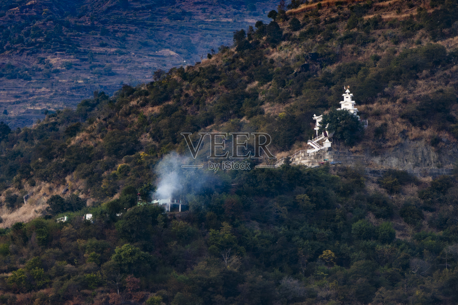 一座白色寺庙坐落在山丘中，设有台阶，四周被绿色植被环绕。附近房屋冒出的白烟为特赫里湖增添了宁静的氛围。照片摄影图片