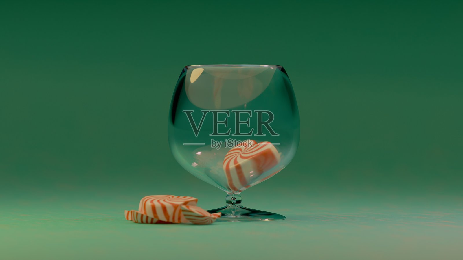 红白相间的心形糖果静静地放在一只空透明玻璃旁边，背景是一片简单的绿色表面。照片摄影图片