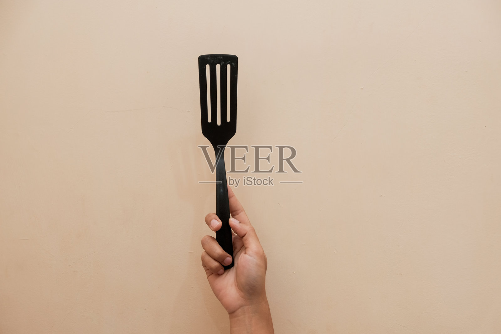 特写镜头：男人手中握着黑色塑料煎铲，背景为奶油色。照片摄影图片