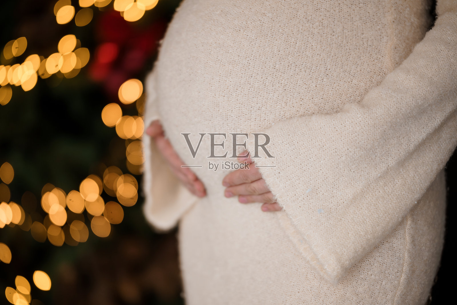 期待中的母亲穿着柔软的毛衣，怀抱着孕肚，背景是节日灯光。照片摄影图片