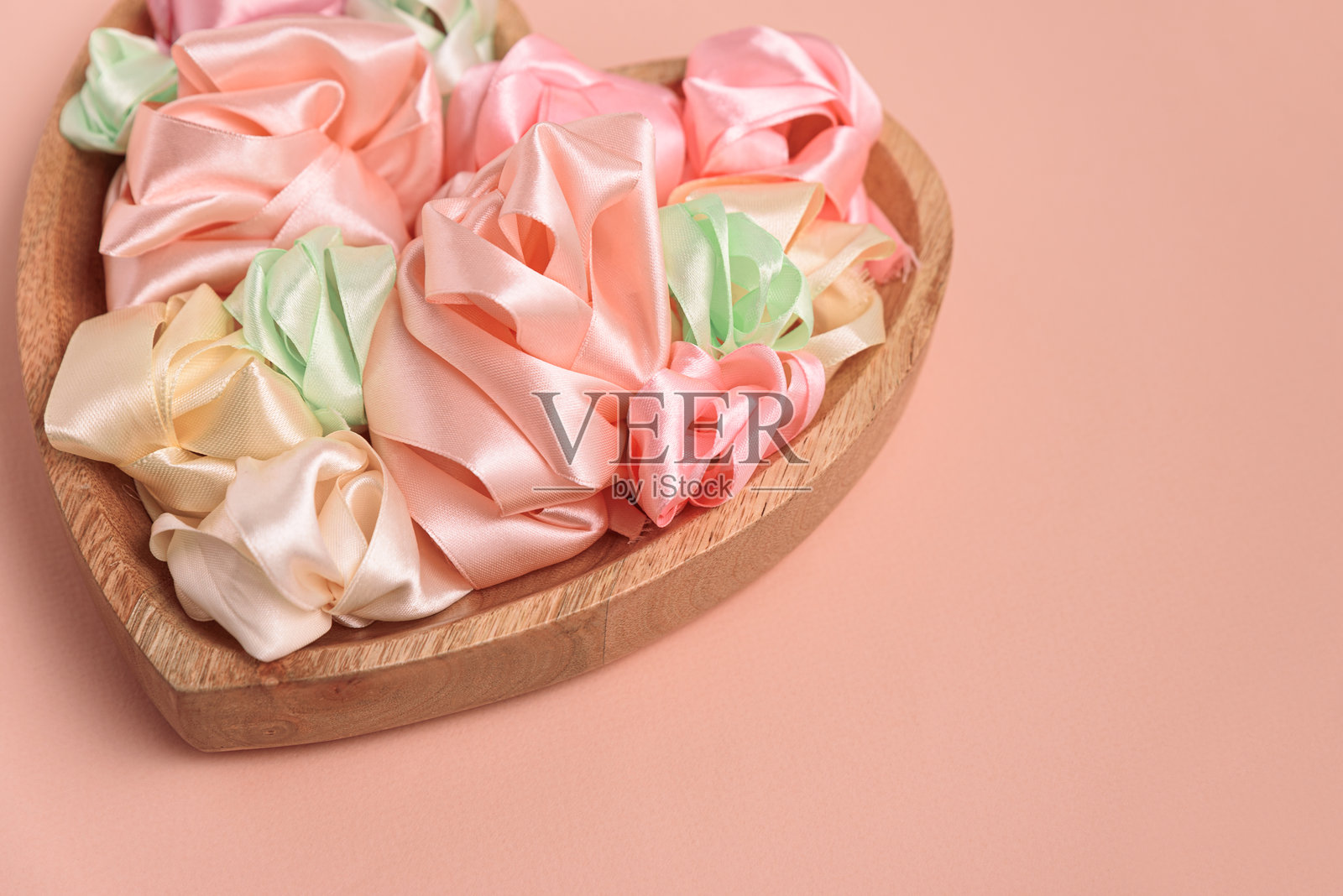 一个心形木碗里装满了柔和色彩的缎带，卷成精致的玫瑰。柔和的粉色背景营造出浪漫而梦幻的氛围。照片摄影图片