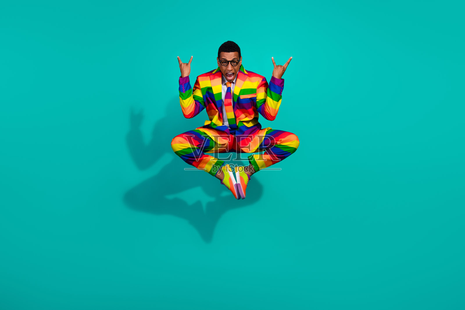 年轻男子穿着鲜艳多彩的西装在青色背景前跳跃，表达快乐、趣味和个性。照片摄影图片