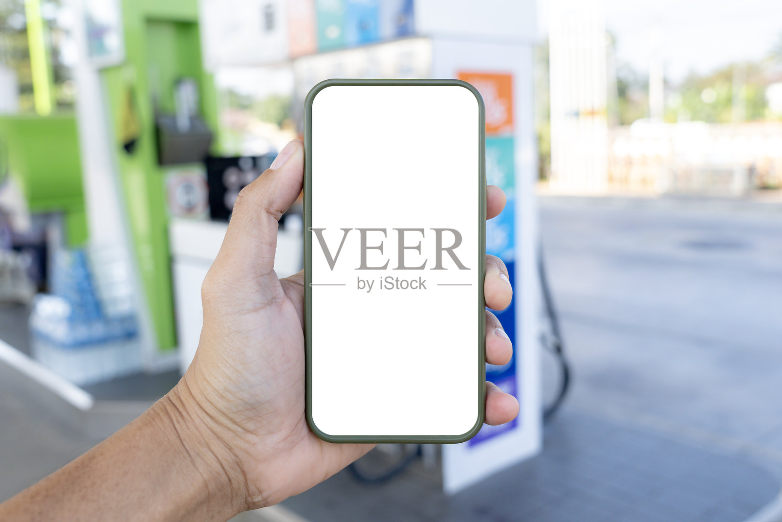 一只手握着一部屏幕空白的智能手机，背景是一个加油站。照片摄影图片