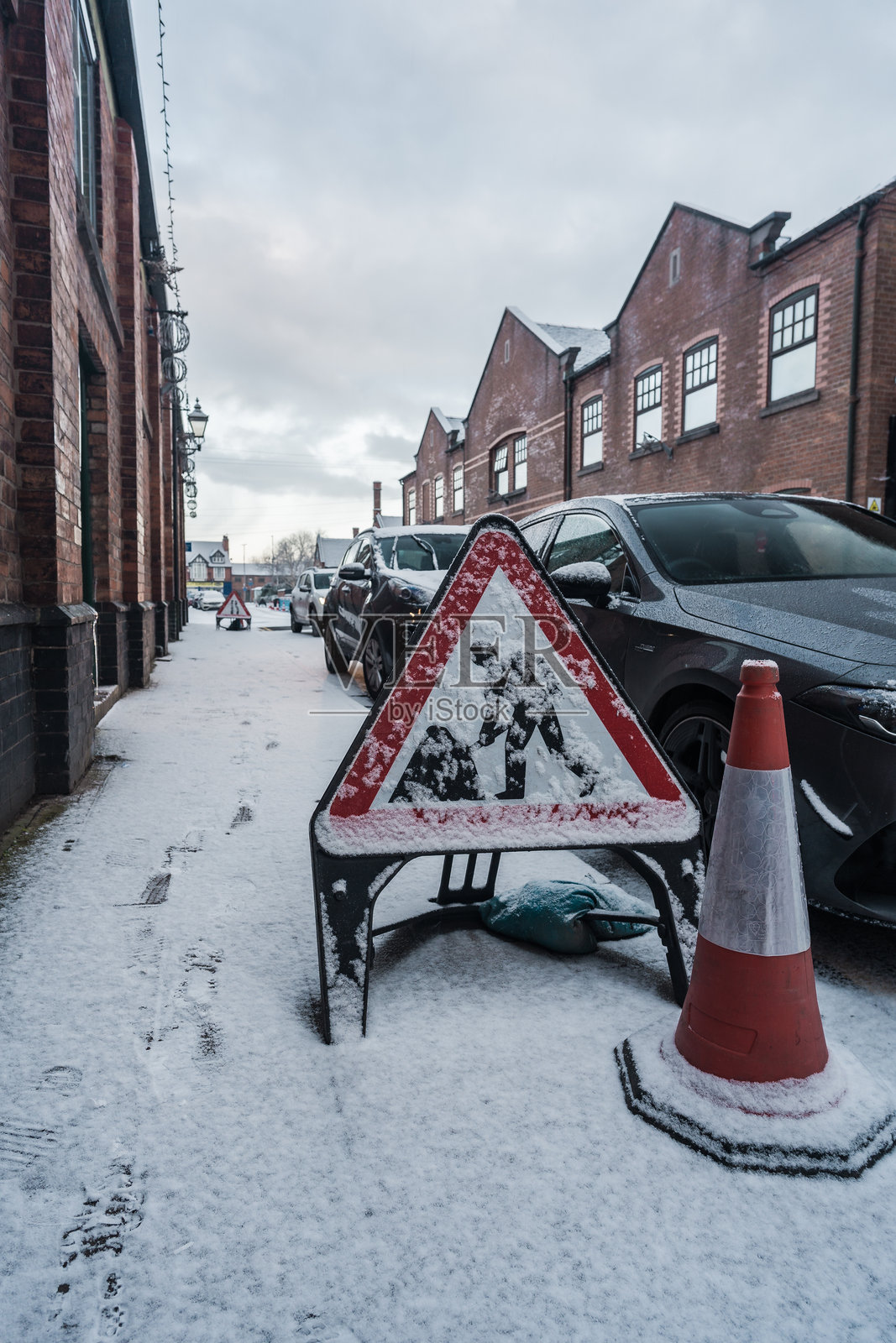 一块被雪覆盖的施工标志和交通锥放置在人行道上，背景中有一辆车。照片摄影图片