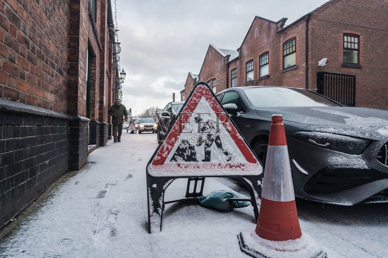 一块被雪覆盖的施工标志和交通锥放置在人行道上，背景中有一辆汽车。照片摄影图片