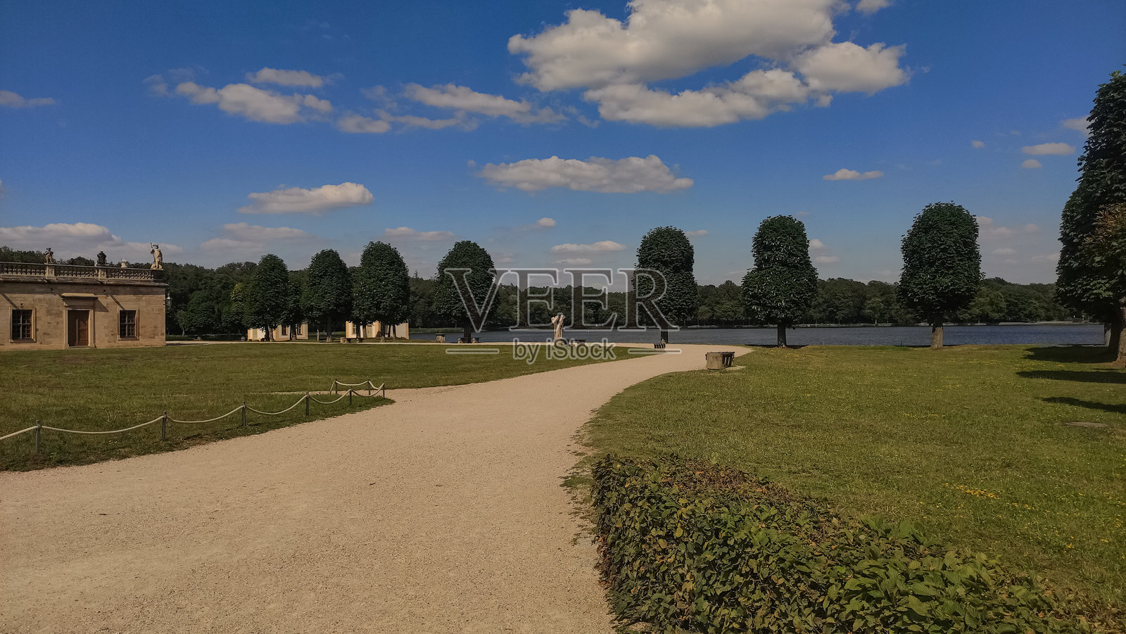 德国莫里茨堡 - 2024年6月25日：一条碎石小路蜿蜒穿过草地，通向湖泊和树木，阳光明媚，靠近城堡。照片摄影图片