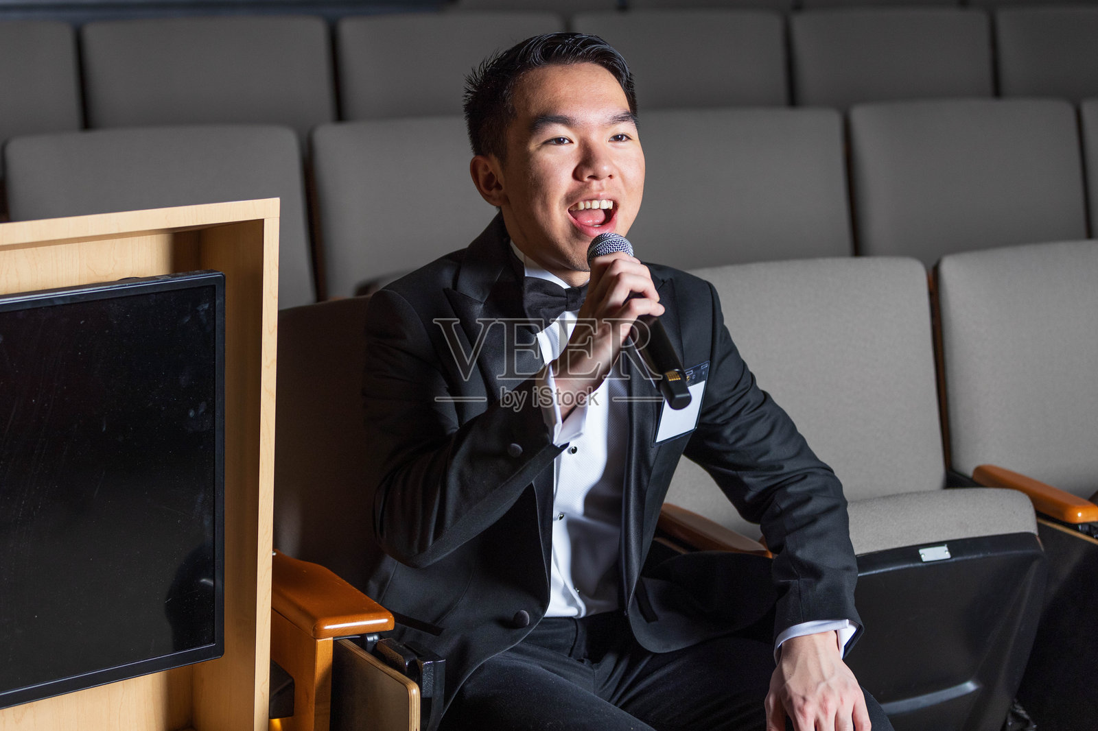 一位身穿晚礼服的男士坐在礼堂里，手里拿着麦克风。照片摄影图片