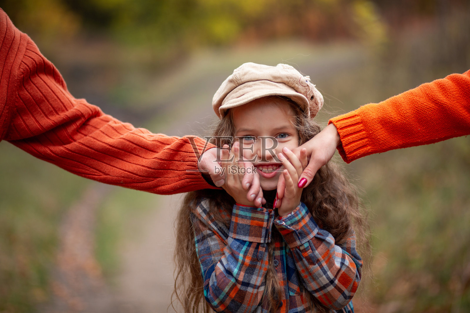 一个七岁金发女孩，长发披肩，戴着帽子，穿着格子衬衫，在秋天的公园里牵着爸爸妈妈的手。照片摄影图片