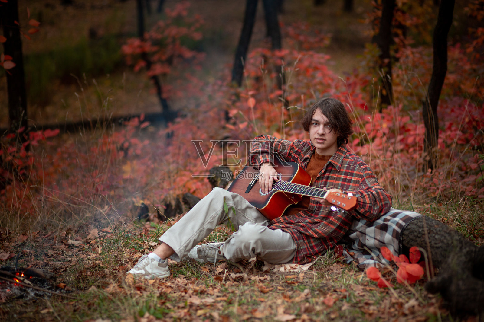 一个穿着格子衬衫的青少年坐在秋季公园的毯子上，边弹吉他边靠近篝火。照片摄影图片