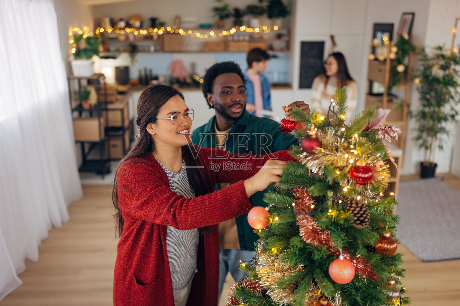 微笑的亚洲女性与她的黑人朋友一起装饰圣诞树。他们在客厅里享受新年和圣诞节的欢乐气氛。照片摄影图片