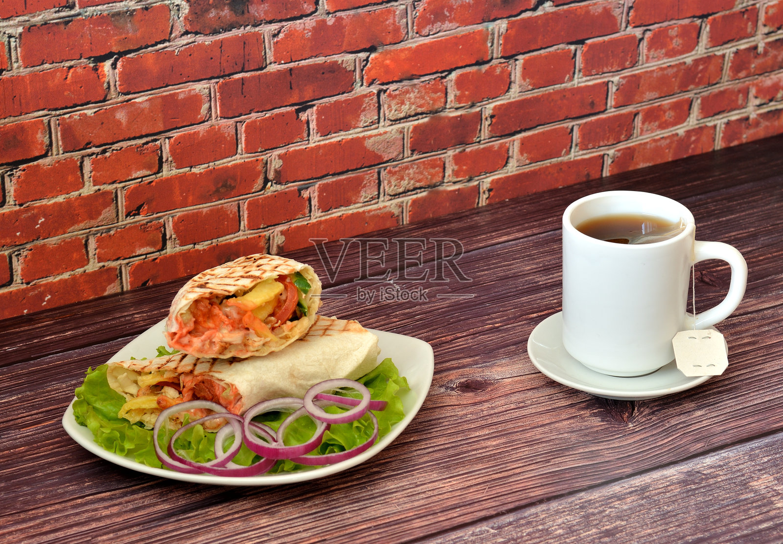 多汁的肉沙威玛放在生菜叶上，配有红洋葱圈和一杯热袋装茶，摆放在木桌上。照片摄影图片