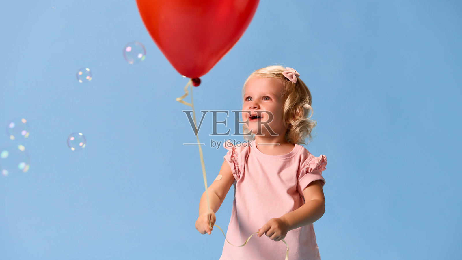 可爱的小女孩兴奋地伸手去抓肥皂泡，穿着粉色衣服，手臂上有绷带，背景是蓝色的摄影棚。照片摄影图片