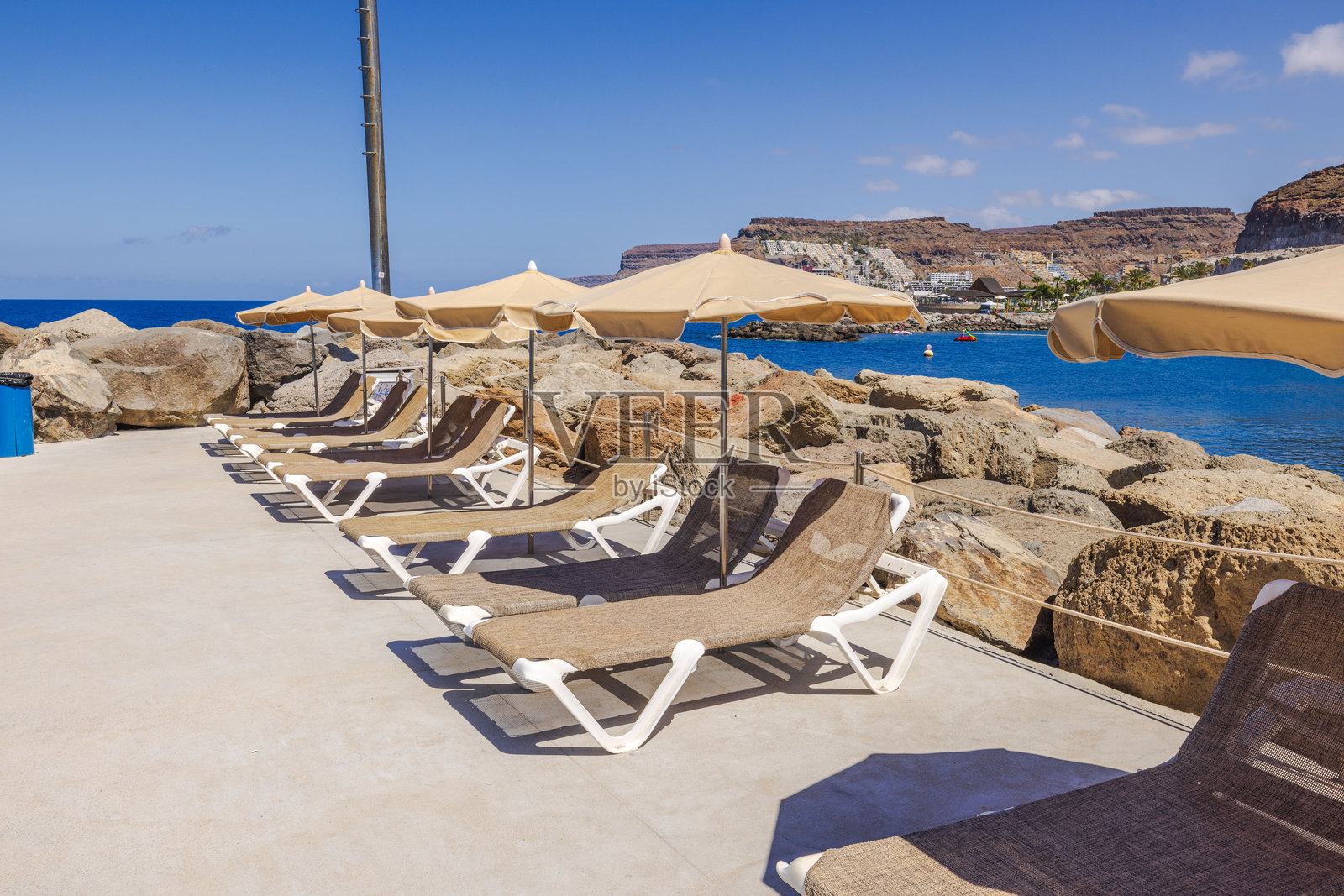 一排空荡荡的日光浴椅在米色伞下，面朝大西洋，背景是岩石海岸线和悬崖。西班牙，拉斯帕尔马斯，格兰 Canaria。照片摄影图片