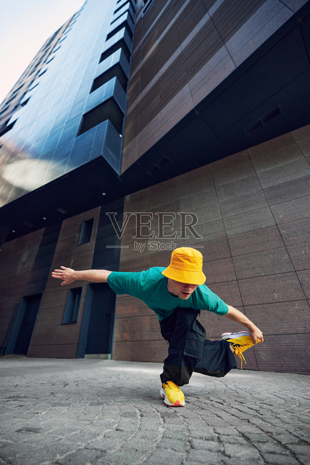 年轻时尚男士穿着色彩鲜艳的服装，在高大现代建筑的城市背景下摆出霹雳舞姿势。照片摄影图片