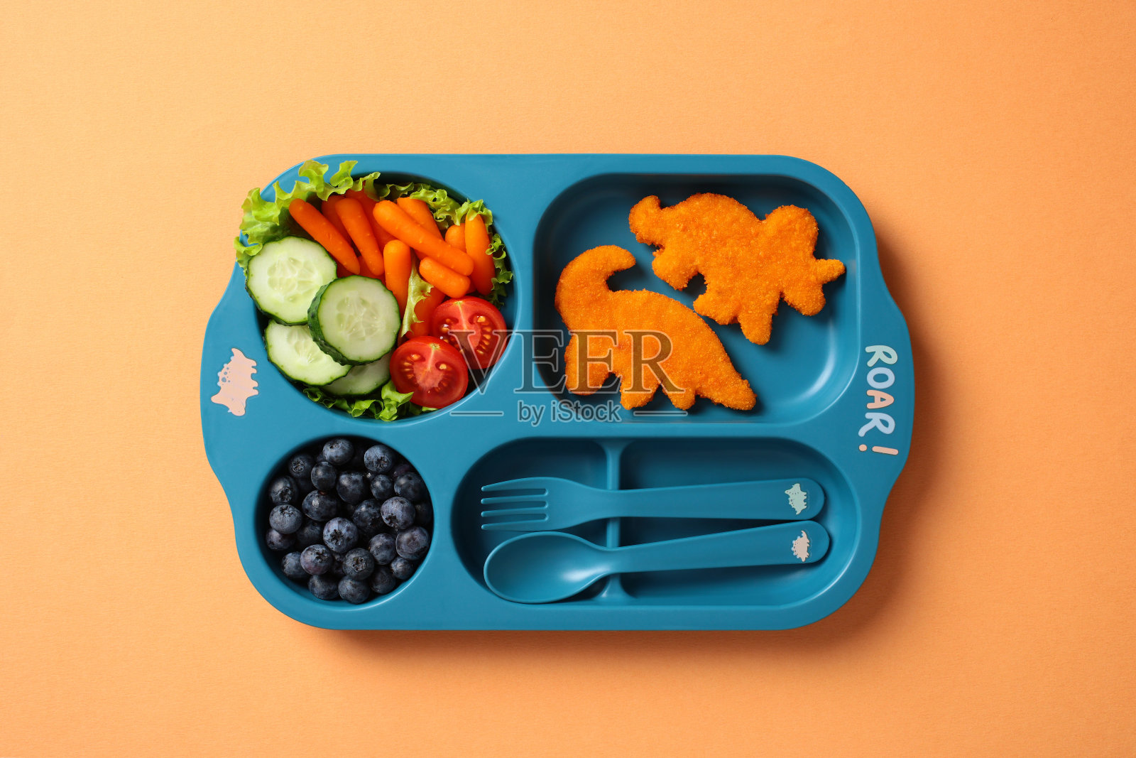 蓝色儿童午餐托盘，里面有恐龙形状的鸡块、新鲜蔬菜、蓝莓和餐具，背景为橙色。适合儿童的有趣健康餐食展示。照片摄影图片