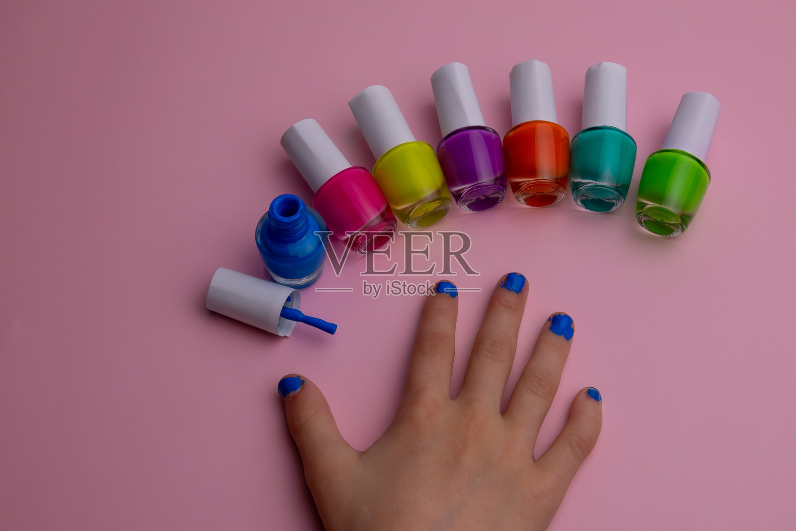 五颜六色的指甲油瓶半圆形排列在粉色表面上，一只手展示着蓝色指甲油。照片摄影图片