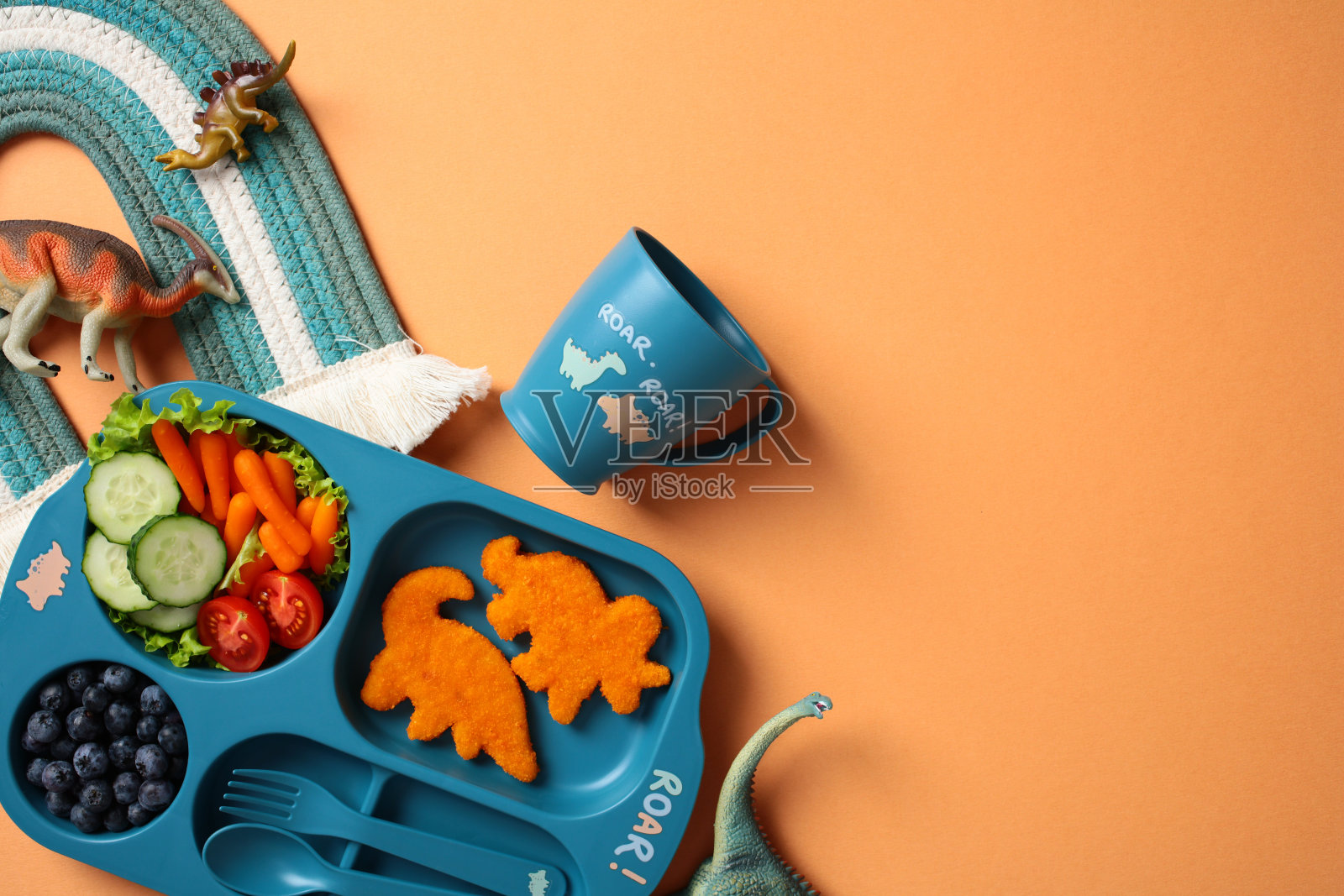 儿童餐具套装，包含恐龙形状的鱼块、新鲜蔬菜和蓝莓，放在蓝色托盘上，配有相配的杯子和玩具恐龙，背景为橙色。照片摄影图片