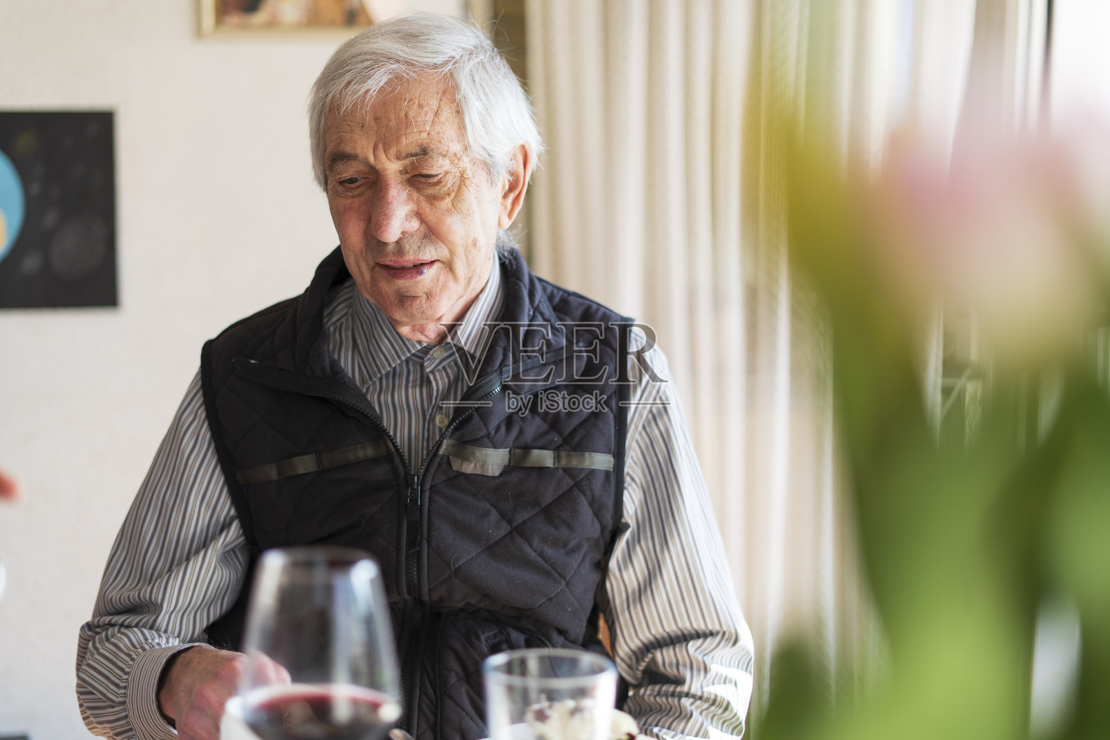 一位穿着衬衫和背心的老人坐在节日的餐桌旁，前景中有一杯酒和一束花。老年快乐的概念照片摄影图片