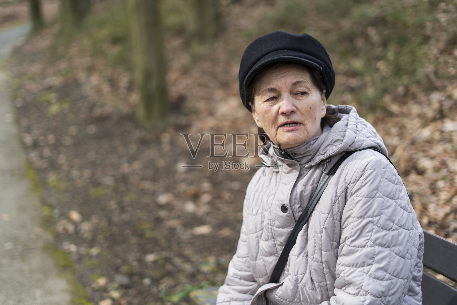 一位穿着夹克和帽子的老年妇女独自坐在公园的长椅上。老年孤独的概念照片摄影图片