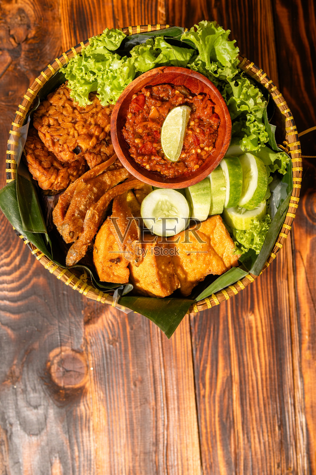 用编织竹篮盛装的传统印尼美食拼盘，包含炸鱼、豆腐、黄瓜、生菜和装在木碗里的沙爹辣椒酱，摆放在香蕉叶上。照片摄影图片