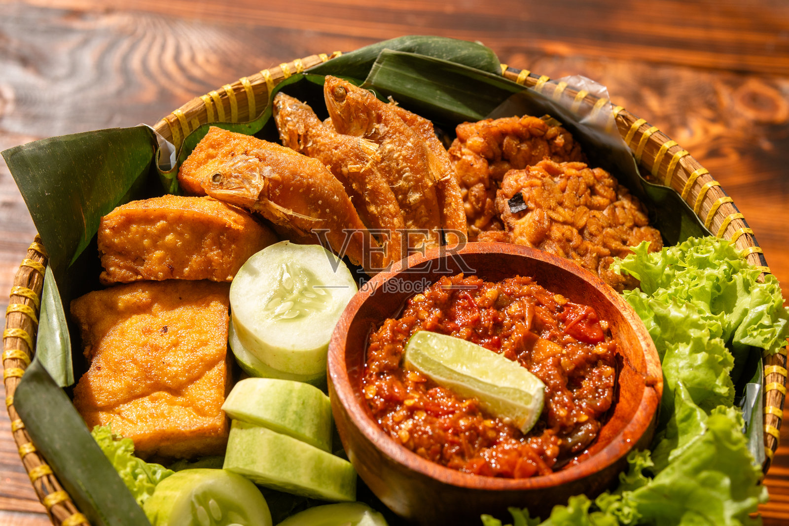 传统印尼美食拼盘，盛放在编织的竹篮中，包含炸鱼、豆腐、黄瓜、生菜和装在木碗里的沙爹辣椒酱，摆放在香蕉叶上。照片摄影图片